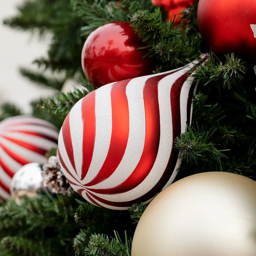 A close-up of a red and white striped teardrop Christmas ornament hanging on a green Christmas tree, surrounded by other red and gold ornaments.
