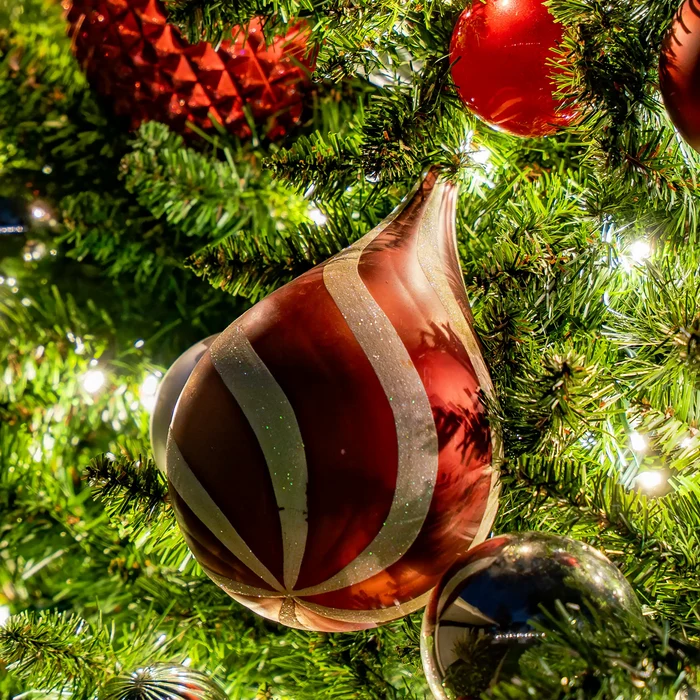 A close-up of a glossy holiday ornament hanging on a green Christmas tree with warm string lights. The ornament is teardrop-shaped with vertical metallic silver stripes on a deep red-brown glossy background.