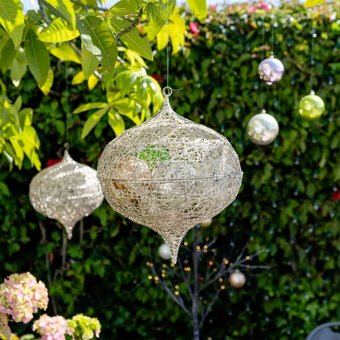 A decorative hanging ornament made of silver-toned mesh or netting, suspended outdoors in front of a green hedge with other ornaments visible in the background.