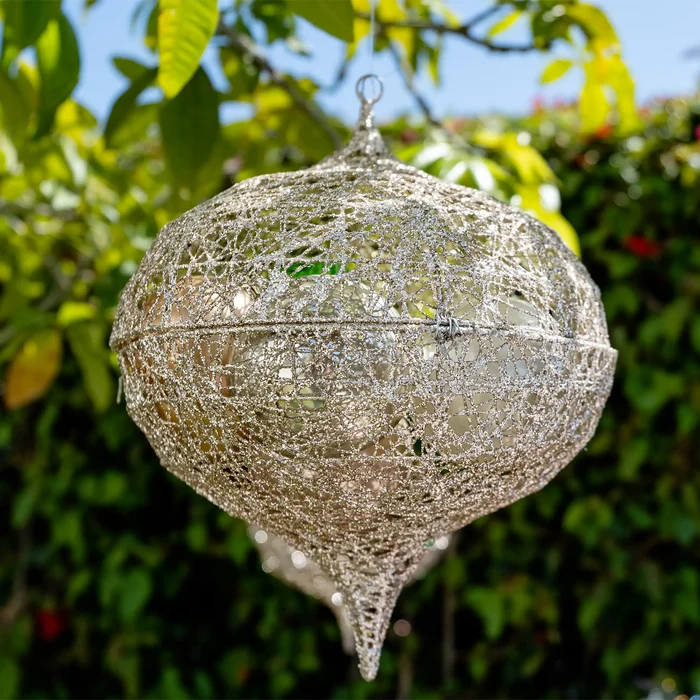 A spherical hanging decorative light made of silver-toned metal mesh and wire, suspended outdoors against a blurred background of green leaves and blue sky.