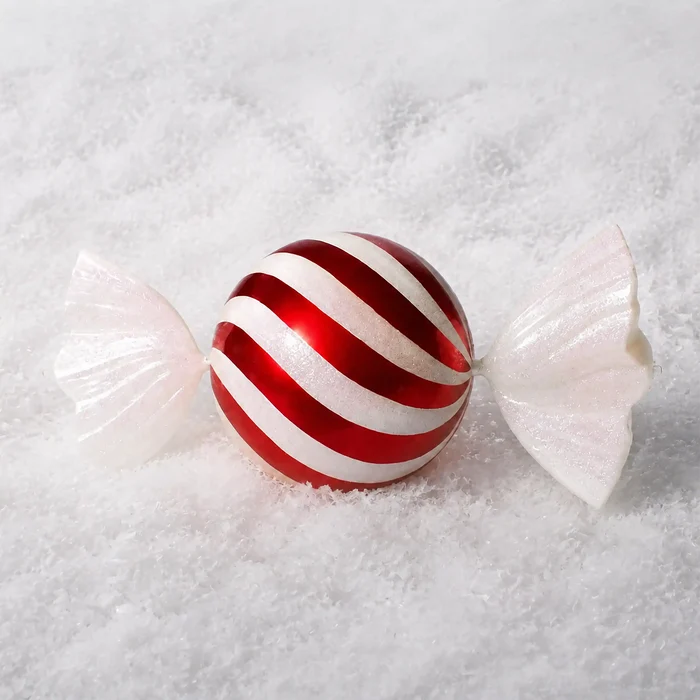 A glossy red and white striped candy ornament or decorative candy with white glittery wings, resting on a textured white snowy surface.