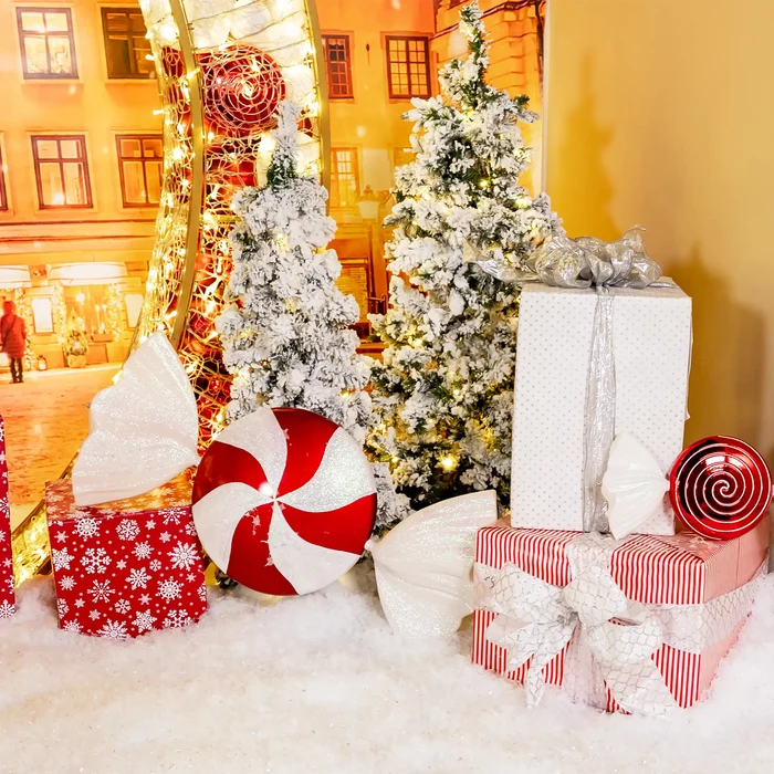 A festive Christmas gift display with wrapped presents, a flocked Christmas tree, a real Christmas tree with warm lights, and large candy-themed ornaments on a white faux-snow surface.
