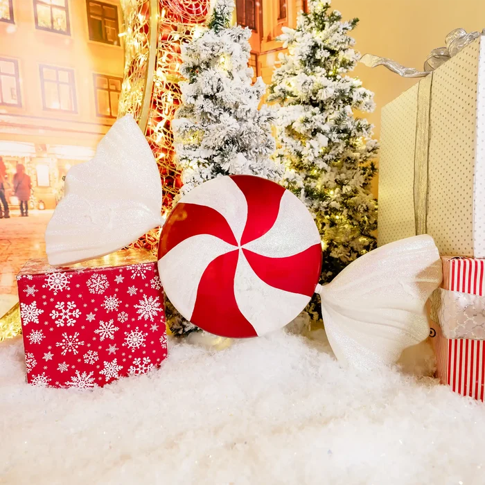 A festive holiday display featuring a large round red and white peppermint candy ornament in the center, flanked by two white angel wings, with wrapped gift boxes and snow-covered Christmas trees in the background.