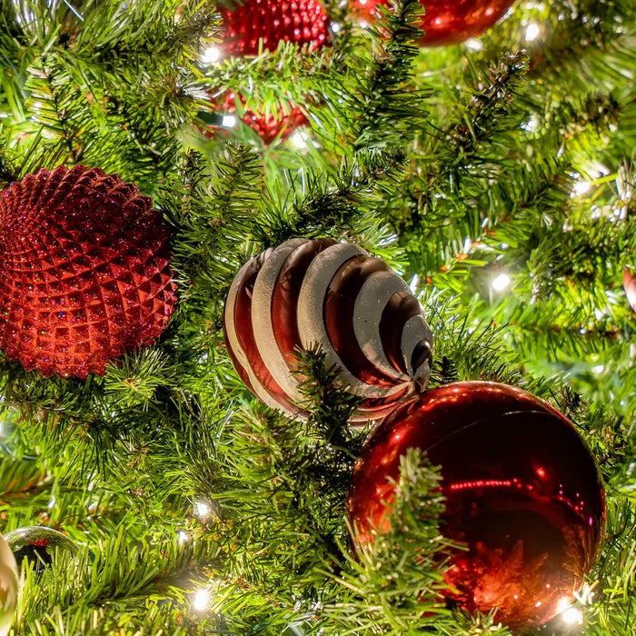 Close-up of a decorated Christmas tree with green branches, warm white lights, and red ornaments, including a large glossy red ball and a striped red-and-white ornament.