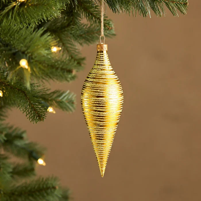 A gold ribbed Christmas ornament shaped like a teardrop or cone, hanging from a green Christmas tree branch with warm string lights in the background.