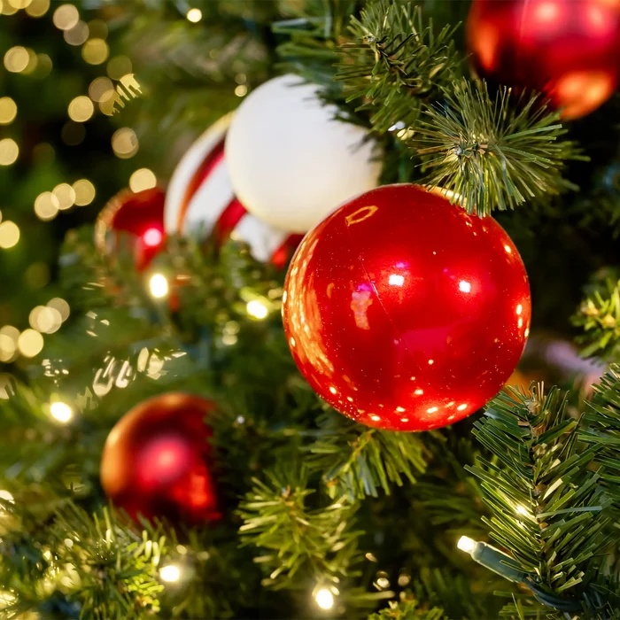A close-up of Dekra-Lite's Candy Apple red Christmas tree ornaments hanging on green artificial tree branches with warm white bokeh lights in the background.
