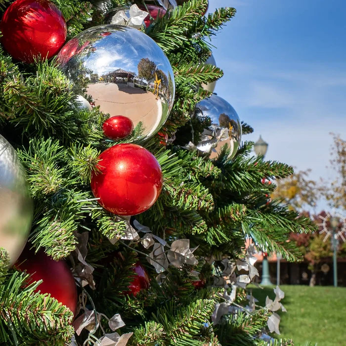 Red and silver Christmas tree ornaments hanging on a green artificial Christmas tree outdoors, with a blue sky and park background visible.