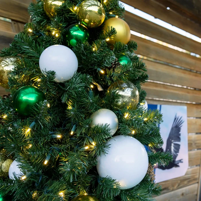 A close-up of a Christmas tree decorated with shiny green and gold ornaments, matte white baubles, and warm yellow string lights, set against a rustic wooden wall.