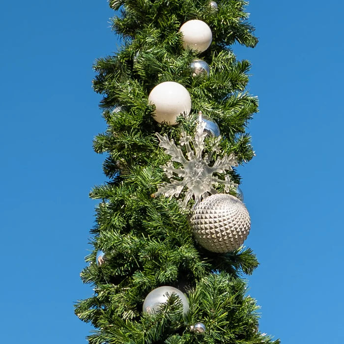 A close-up of a green artificial Christmas tree branch decorated with white, silver, and clear ornaments against a solid blue sky background.