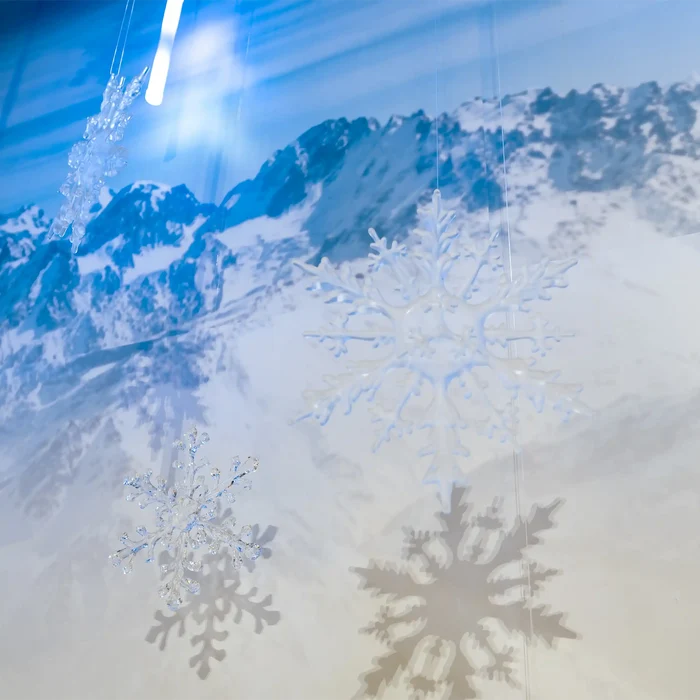 Three translucent white snowflake ornaments hanging from thin wires against a snowy mountain landscape backdrop, with blue and gray shadows cast on the surface below.