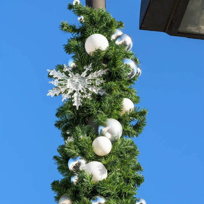 A close-up of a green artificial Christmas tree garland decorated with white and silver ornaments, including a large white snowflake, hanging against a clear blue sky.