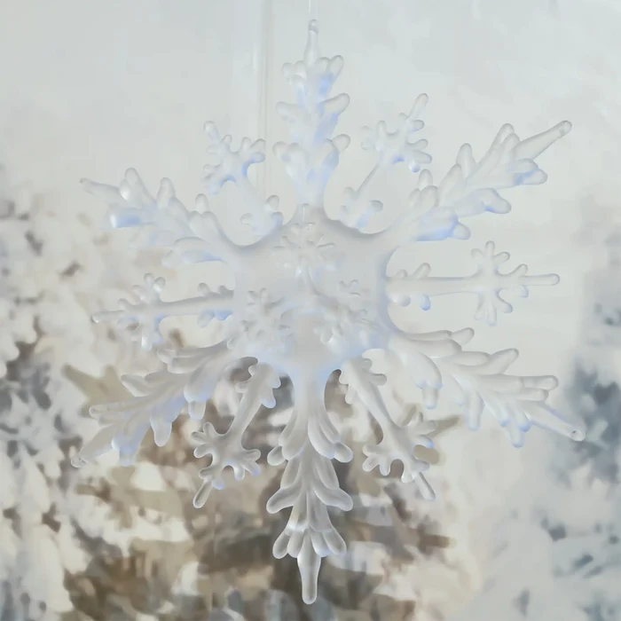 A translucent white snowflake ornament hanging against a softly blurred winter background with frosty branches.