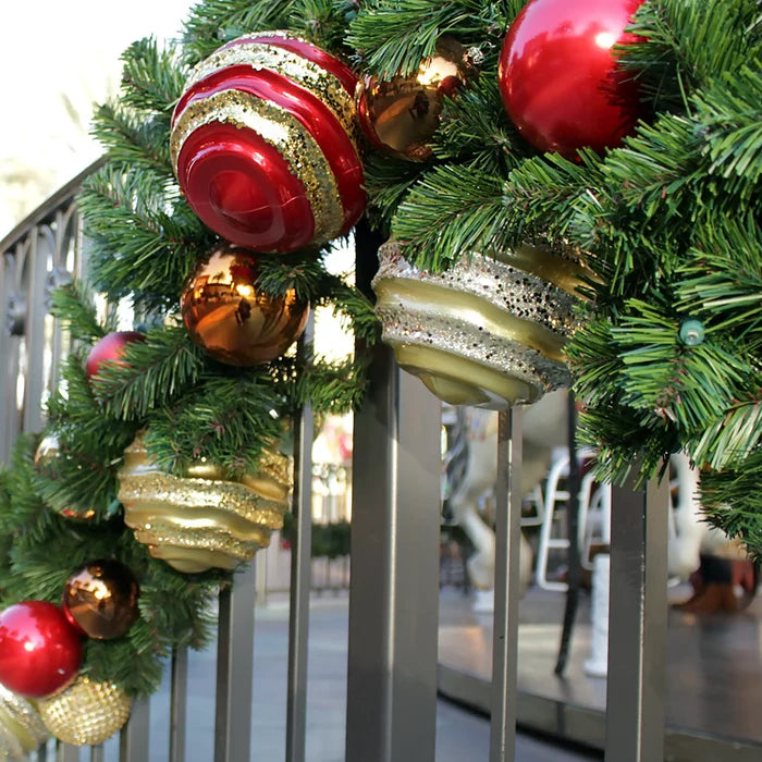 A close-up of a green Christmas garland draped over a gray metal railing, decorated with shiny red and gold ornaments and metallic gold and bronze baubles.