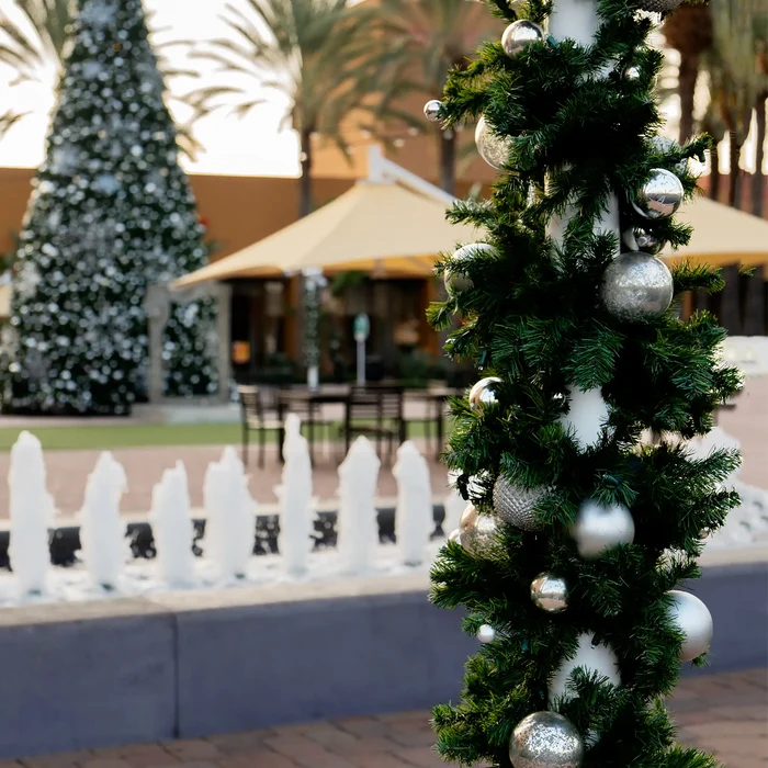 A decorated Christmas tree trunk in an outdoor patio setting, wrapped in dark green artificial pine needles and adorned with shiny silver and white ornaments, with a blurred festive background.